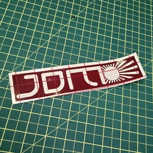May include: A rectangular sticker featuring the word "JDM" in white, a stylized font, and a rising sun design in white and red. The sticker is on a green cutting mat with grid lines.