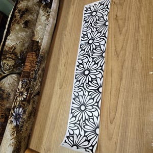 K&ouml;nnte beinhalten: Ein langer, schmaler, wei&szlig;er Aufkleber mit einem sich wiederholenden schwarzen Blumenmuster. Der Aufkleber ist teilweise von einer hellbraunen Holzoberfl&auml;che abgezogen. Das Blumenmuster zeigt stilisierte, g&auml;nsebl&uuml;mchenartige Blumen.
