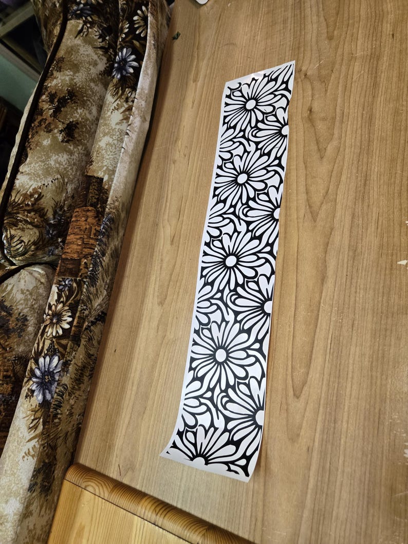 K&ouml;nnte beinhalten: Ein langer, schmaler, wei&szlig;er Aufkleber mit einem sich wiederholenden schwarzen Blumenmuster. Der Aufkleber ist teilweise von einer hellbraunen Holzoberfl&auml;che abgezogen. Das Blumenmuster zeigt stilisierte, g&auml;nsebl&uuml;mchenartige Blumen.
