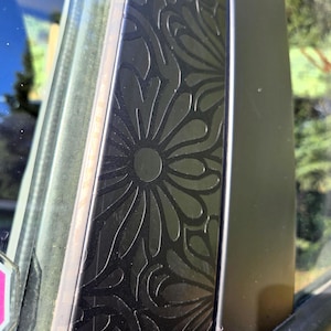 K&ouml;nnte beinhalten: Nahaufnahme einer schwarzen Dekorplatte mit floralem Muster. Das Design zeigt stilisierte Blumen und wirbelnde Bl&auml;tter. Die Platte ist neben einer Autoscheibe montiert, wobei ein Blick auf die Au&szlig;enwelt durch das Glas sichtbar ist.