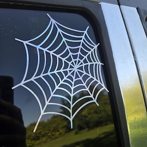 May include: A holographic spiderweb sticker applied to a car window. The web design is white with an iridescent sheen, reflecting various colours. The sticker is on dark tinted glass, with a view of a green lawn visible through the window.