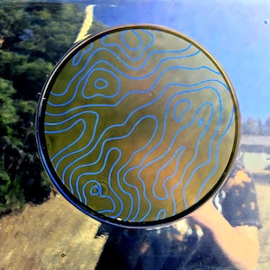 May include: A circular, dark green, wooden coaster with a blue, wavy design. The coaster is on a blue surface.