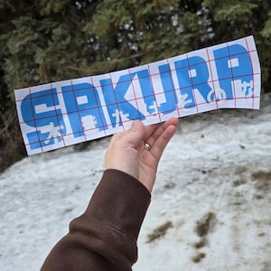 May include: A blue vinyl sticker with the word "SAKURP" in white letters. The sticker is on a gridded backing sheet.