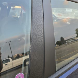 K&ouml;nnte beinhalten: Nahaufnahme eines Autofensters mit einem floralen Muster auf dem Rahmen. Das Fenster ist get&ouml;nt, und ein rosa-wei&szlig;er Aufkleber ist sichtbar. Das &Auml;u&szlig;ere des Autos ist blau.