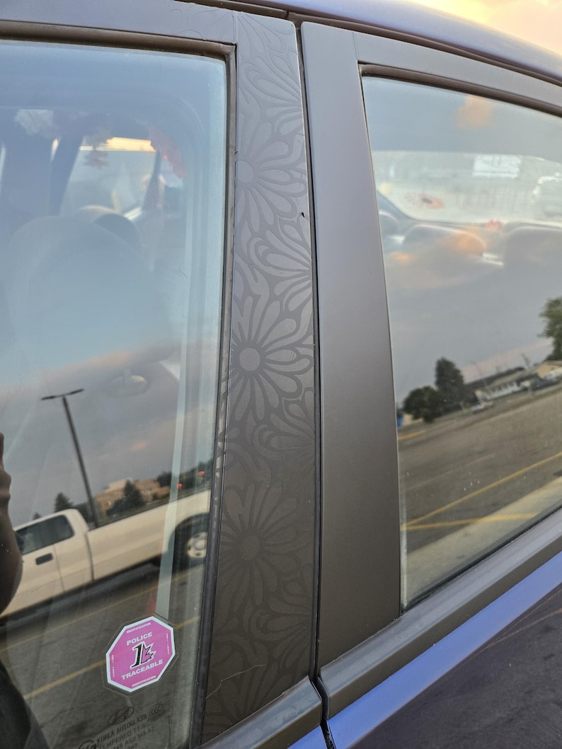 K&ouml;nnte beinhalten: Nahaufnahme eines Autofensters mit einem floralen Muster auf dem Rahmen. Das Fenster ist get&ouml;nt, und ein rosa-wei&szlig;er Aufkleber ist sichtbar. Das &Auml;u&szlig;ere des Autos ist blau.