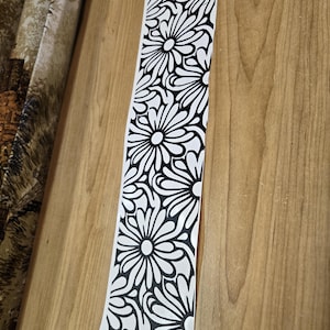 K&ouml;nnte beinhalten: Ein langer, schmaler Streifen aus wei&szlig;em Material mit einem sich wiederholenden schwarzen Blumenmuster. Das Design zeigt gro&szlig;e, stilisierte Blumen mit detaillierten Bl&uuml;tenbl&auml;ttern, die eine k&uuml;hne, grafische &Auml;sthetik erzeugen. Der Streifen ist teilweise abgerollt.