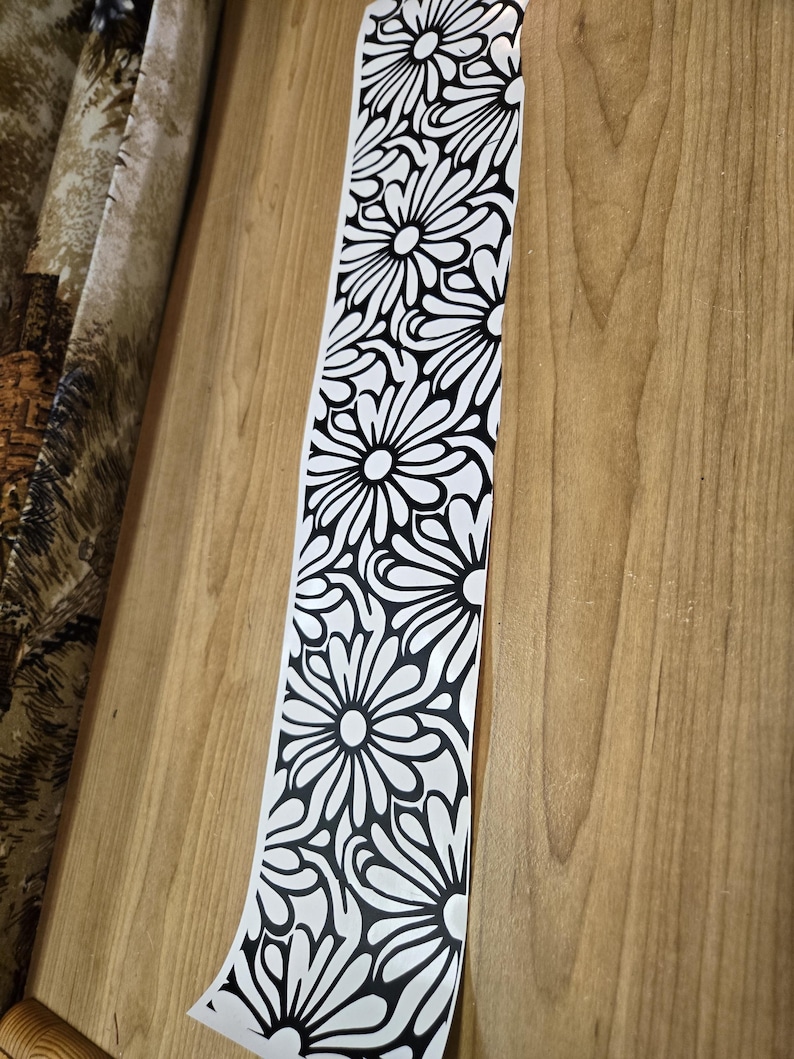 K&ouml;nnte beinhalten: Ein langer, schmaler Streifen aus wei&szlig;em Material mit einem sich wiederholenden schwarzen Blumenmuster. Das Design zeigt gro&szlig;e, stilisierte Blumen mit detaillierten Bl&uuml;tenbl&auml;ttern, die eine k&uuml;hne, grafische &Auml;sthetik erzeugen. Der Streifen ist teilweise abgerollt.