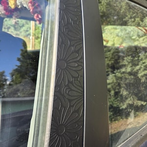K&ouml;nnte beinhalten: Nahaufnahme eines Autot&uuml;rrahmens mit einem dekorativen Blumenmuster. Der schwarze Rahmen hat ein gepr&auml;gtes Design aus sich wiederholenden stilisierten Blumen. Der Fensterrahmen ist sichtbar, mit einem Blick auf die Au&szlig;enwelt, einschlie&szlig;lich B&auml;umen und Himmel.