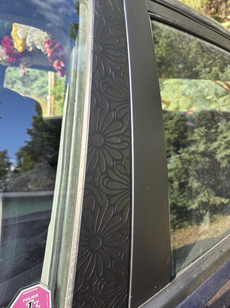 K&ouml;nnte beinhalten: Nahaufnahme eines Autot&uuml;rrahmens mit einem dekorativen Blumenmuster. Der schwarze Rahmen hat ein gepr&auml;gtes Design aus sich wiederholenden stilisierten Blumen. Der Fensterrahmen ist sichtbar, mit einem Blick auf die Au&szlig;enwelt, einschlie&szlig;lich B&auml;umen und Himmel.