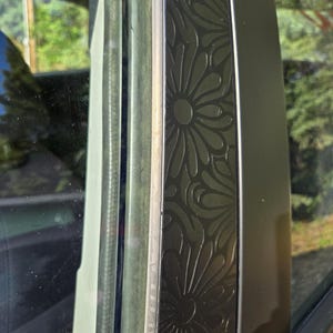 K&ouml;nnte beinhalten: Nahaufnahme eines Autofensterrahmens mit einem dekorativen Blumenmuster. Der Rahmen ist dunkel, m&ouml;glicherweise schwarz, mit einem sich wiederholenden Design aus stilisierten Blumen. Das Glas ist sichtbar und reflektiert die Au&szlig;enumgebung.