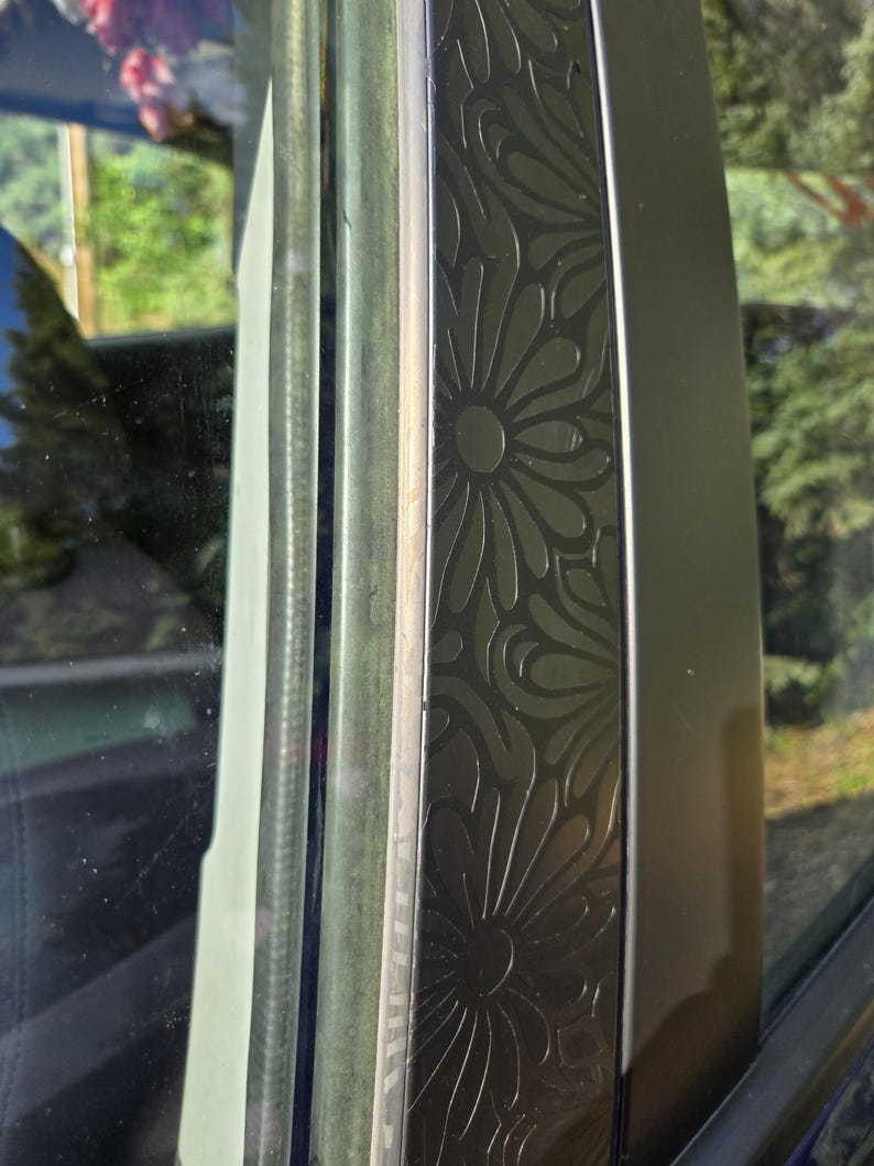 K&ouml;nnte beinhalten: Nahaufnahme eines Autofensterrahmens mit einem dekorativen Blumenmuster. Der Rahmen ist dunkel, m&ouml;glicherweise schwarz, mit einem sich wiederholenden Design aus stilisierten Blumen. Das Glas ist sichtbar und reflektiert die Au&szlig;enumgebung.