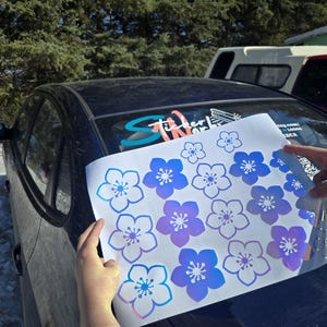 May include: A sheet of flower-shaped stickers in shades of blue and purple is held over a dark blue car. The stickers have a gradient effect, with a light blue outline and a darker centre. The background includes trees and a vehicle.
