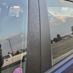 K&ouml;nnte beinhalten: Nahaufnahme eines Autot&uuml;rrahmens mit floralem Muster. Der dunkelgraue Rahmen hat ein sich wiederholendes, g&auml;nsebl&uuml;mchenartiges Design. Das Fenster des Autos spiegelt den Himmel und einen wei&szlig;en Pickup-Truck wider. Ein rosa-wei&szlig;er Aufkleber ist sichtbar.