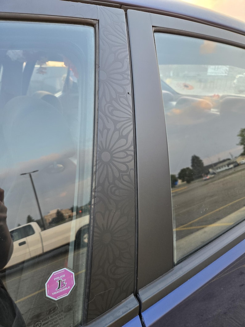 K&ouml;nnte beinhalten: Nahaufnahme eines Autot&uuml;rrahmens mit floralem Muster. Der dunkelgraue Rahmen hat ein sich wiederholendes, g&auml;nsebl&uuml;mchenartiges Design. Das Fenster des Autos spiegelt den Himmel und einen wei&szlig;en Pickup-Truck wider. Ein rosa-wei&szlig;er Aufkleber ist sichtbar.