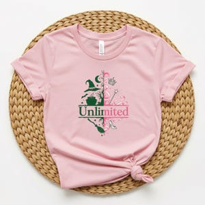 May include: A light pink t-shirt with rolled-up sleeves. The shirt features a split design with a witch hat, cauldron, and broom on one side and a crown, wand, and high heel on the other. The word "Unlimited" is printed in green and pink.