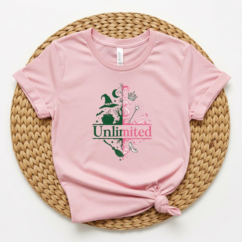 May include: A light pink t-shirt with rolled-up sleeves. The shirt features a split design with a witch hat, cauldron, and broom on one side and a crown, wand, and high heel on the other. The word "Unlimited" is printed in green and pink.