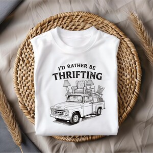 May include: White t-shirt with the text "I'D RATHER BE THRIFTING" in black. The graphic shows a vintage pickup truck loaded with furniture, including lamps, a chair, and other items. The shirt is displayed on a woven basket.