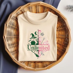 May include: A cream-colored t-shirt with a split design featuring a witch-themed side and a princess-themed side. The witch side includes a hat, cauldron, and moon. The princess side has a crown, wand, and shoe. The word "Unlimited" is printed across the bottom.