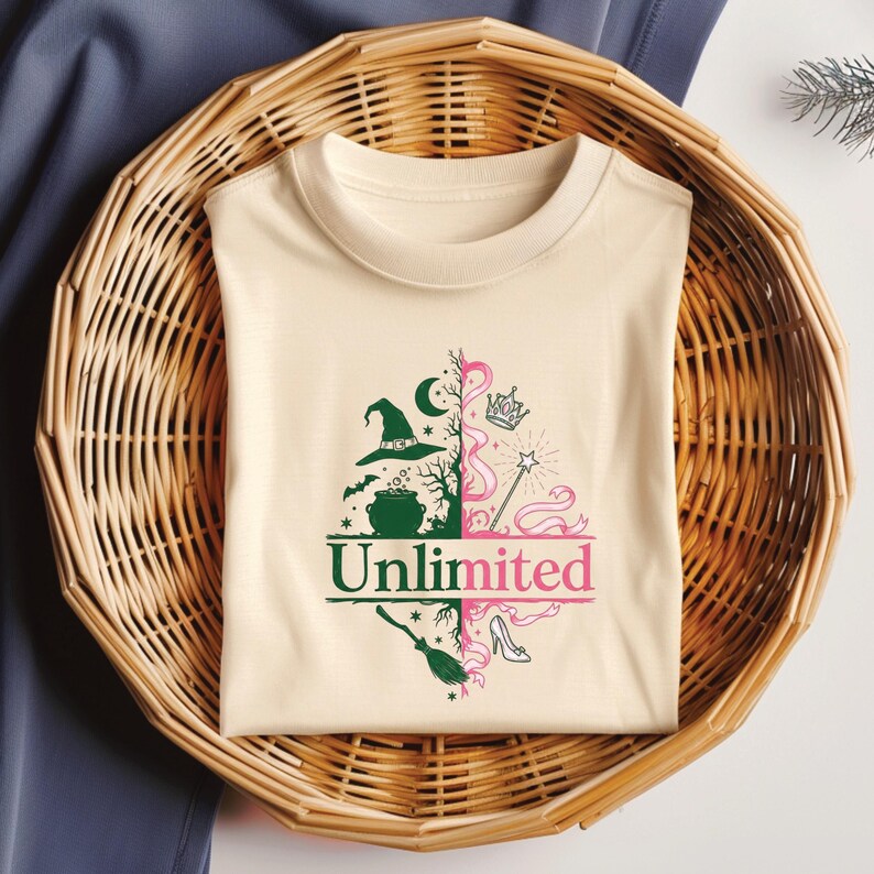 May include: A cream-colored t-shirt with a split design featuring a witch-themed side and a princess-themed side. The witch side includes a hat, cauldron, and moon. The princess side has a crown, wand, and shoe. The word "Unlimited" is printed across the bottom.