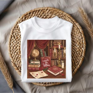 May include: White t-shirt featuring a detailed illustration of a library scene with books, candles, and a globe. The graphic includes the text "I Prefer the Villain" in a decorative font, set against a backdrop of red curtains and bookshelves. The shirt is displayed on a woven basket.