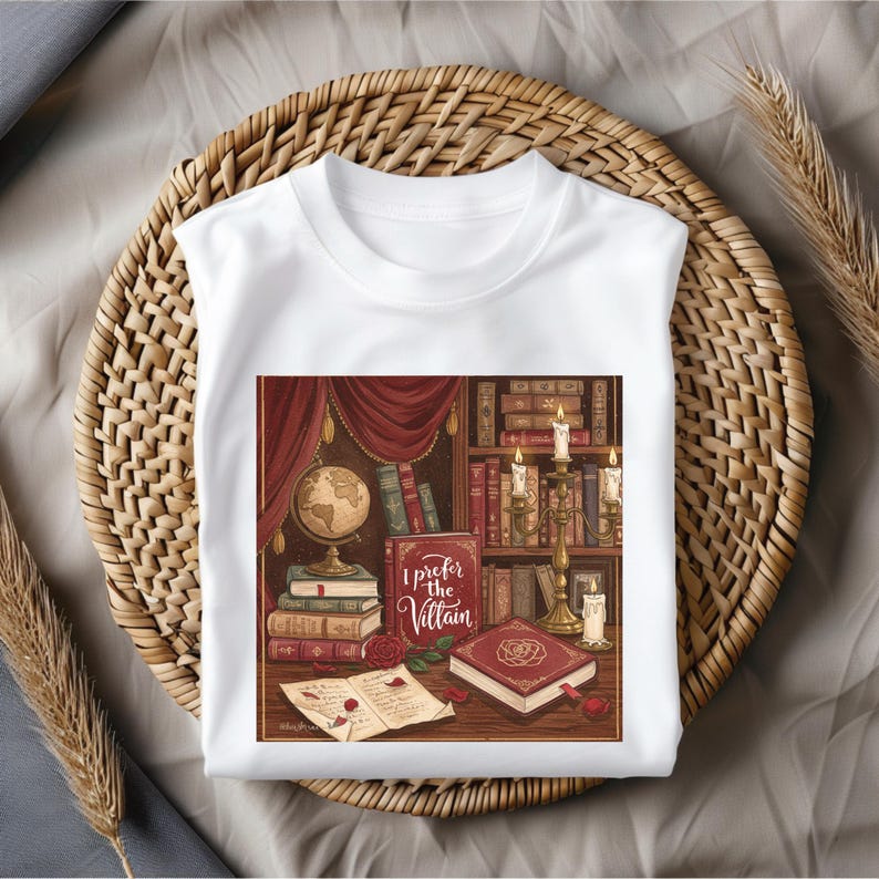 May include: White t-shirt featuring a detailed illustration of a library scene with books, candles, and a globe. The graphic includes the text "I Prefer the Villain" in a decorative font, set against a backdrop of red curtains and bookshelves. The shirt is displayed on a woven basket.