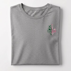 May include: A heather gray t-shirt with a crew neck. The shirt features a small graphic on the left chest with the word "Unlimited" and a design of a witch's hat, broom, and other elements in green and pink.