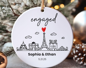 May include: A round white ceramic ornament hanging from a gold ribbon. The ornament features the word engaged in black script, a red heart above a black line drawing of the Paris skyline, and the text Sophia & Ethan with the date 5.21.25.