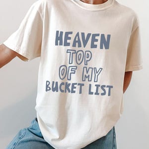 以下が含まれることがあります： クリーム色のTシャツに、青い文字で「HEAVEN TOP OF MY BUCKET LIST」と書かれています。柔らかいコットン素材と思われるカジュアルなTシャツが着用されています。全体的にリラックスした快適な雰囲気です。