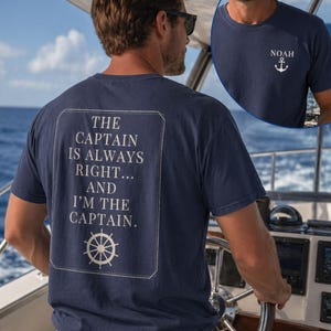 Man wearing a navy nautical captain shirt while steering a boat on the ocean. The back design reads “The Captain Is Always Right… and I’m the Captain” with a ship wheel graphic, and an inset shows the front chest design with the name Noah and an anchor.