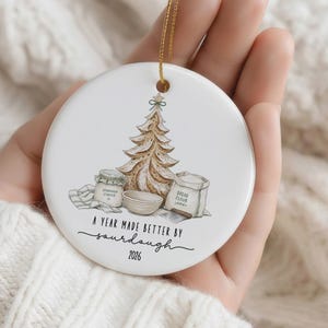 Puede incluir: Un adorno redondo de cer&aacute;mica blanca con una ilustraci&oacute;n marr&oacute;n de un &aacute;rbol de Navidad con forma de pan, un frasco etiquetado Sourdough Starter, un cuenco y una bolsa etiquetada Bread Flour. El texto dice A YEAR MADE BETTER BY sourdough 2026.