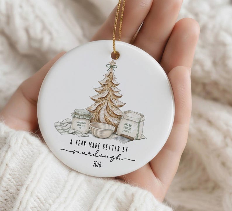 Puede incluir: Un adorno redondo de cer&aacute;mica blanca con una ilustraci&oacute;n marr&oacute;n de un &aacute;rbol de Navidad con forma de pan, un frasco etiquetado Sourdough Starter, un cuenco y una bolsa etiquetada Bread Flour. El texto dice A YEAR MADE BETTER BY sourdough 2026.