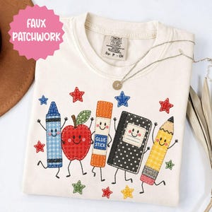 May include: A cream-coloured t-shirt featuring a faux patchwork graphic of a blue gingham crayon, a red gingham apple, an orange glue stick, a black polka-dot notebook, and a yellow gingham pencil, all with smiling faces and small stars.