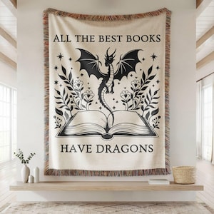 May include: Cream-colored woven throw blanket with a black dragon design and the text "ALL THE BEST BOOKS HAVE DRAGONS." The dragon is perched above an open book, surrounded by floral and star motifs. The blanket has a fringed edge.