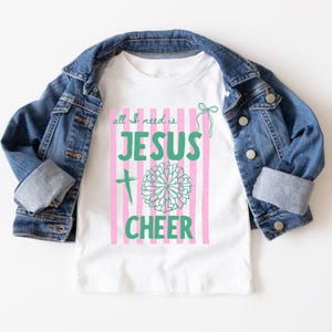 Könnte beinhalten: Ein weißes Kurzarm-T-Shirt mit einem Grafikdruck aus rosa vertikalen Streifen, einem grünen Kreuz, einem grünen Pompon und dem Text "all I need is JESUS CHEER" in Grün, unter einer blauen Jeansjacke mit Metallknöpfen und umgeschlagenen Ärmeln.
