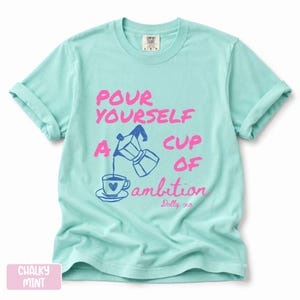 Pu&ograve; includere: Una t-shirt a maniche corte color menta gessato con una grafica blu scuro di una moka che versa il caff&egrave; in una tazzina. Il testo in rosa brillante recita: POUR YOURSELF A CUP OF ambition, con la firma Dolly xo scritta in corsivo sotto.