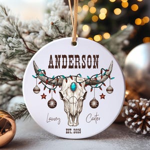 Puede incluir: Un adorno redondo de cerámica blanca con una ilustración de un cráneo de toro decorado con luces navideñas de colores y adornos colgantes. El texto dice ANDERSON arriba, Lainey y Carter en el centro, y EST. 2025 abajo.