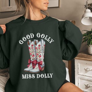 May include: Dark green sweatshirt with the text "GOOD GOLLY MISS DOLLY" in white. The graphic features a pair of white cowboy boots with red floral designs and red bows.