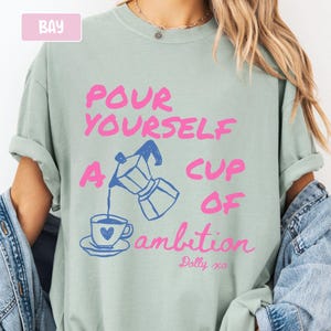 Pu&ograve; includere: Una t-shirt verde salvia con il testo rosa "POUR YOURSELF A CUP OF ambition" e un disegno blu di una caffettiera moka che versa caff&egrave; in una tazza con un cuore. Il testo "Dolly xo" &egrave; stampato in rosa sotto il disegno.