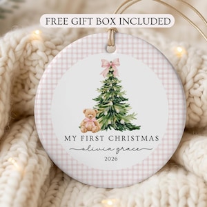 Puede incluir: Un adorno redondo de cerámica con un borde de cuadros vichy en rosa claro y blanco. El centro presenta una ilustración en acuarela de un árbol de Navidad verde con un lazo rosa, un osito de peluche y el texto: MY FIRST CHRISTMAS, olivia grace, 2026.