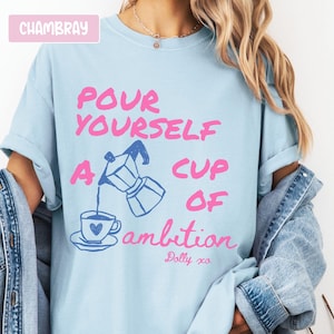 Pu&ograve; includere: Una t-shirt azzurro chiaro con il testo rosa "POUR YOURSELF A CUP OF" e il testo blu "ambition", con un disegno di una moka che versa caff&egrave; in una tazza con un cuore. La maglietta riporta anche le scritte "Dolly xo" e "CHAMBRAY".