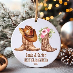 Puede incluir: Un adorno redondo de cerámica blanca con un sombrero de vaquero marrón sobre una bota de vaquero marrón, junto a una segunda bota decorada con flores rosas. El texto dice Lanie & Carter ENGAGED 2025 en letras de color marrón.