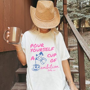 Pu&ograve; includere: Una t-shirt bianca a maniche corte con una moka blu che versa caff&egrave; in una tazza con un cuore. Il testo recita: POUR YOURSELF A CUP OF ambition Dolly xo in rosa e blu. Sono visibili anche un cappello da cowboy in paglia e una tazza marrone.
