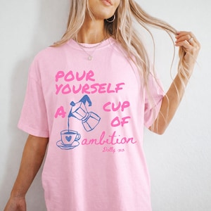 Pu&ograve; includere: Una t-shirt a maniche corte rosa chiaro con una grafica blu di una caffettiera moka che versa il caff&egrave; in una tazza. Il testo recita: POUR YOURSELF A CUP OF ambition, con la firma Dolly xo in basso. Il design &egrave; stampato in rosa e blu.