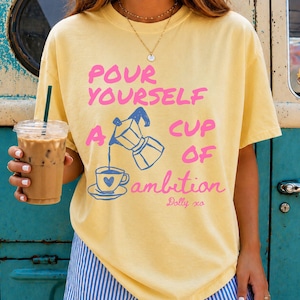 Woman modeling a pastel yellow graphic T-shirt featuring pink and blue lettering that reads “Pour Yourself a Cup of Ambition” with a coffee pot and cup illustration, while holding an iced coffee in front of a vintage turquoise vehicle.