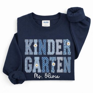 May include: A navy blue sweatshirt featuring the words "KINDER GARDEN" in patchwork lettering, decorated with daisies. The name "Ms. Olivia" is printed below. The sweatshirt is made of a soft material.