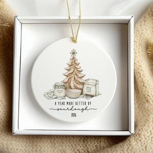 Puede incluir: Un adorno redondo de cer&aacute;mica blanca con una ilustraci&oacute;n en acuarela de un &aacute;rbol de Navidad de pan de masa madre, un frasco, un cuenco y una bolsa de harina. El texto dice A YEAR MADE BETTER BY sourdough 2026.