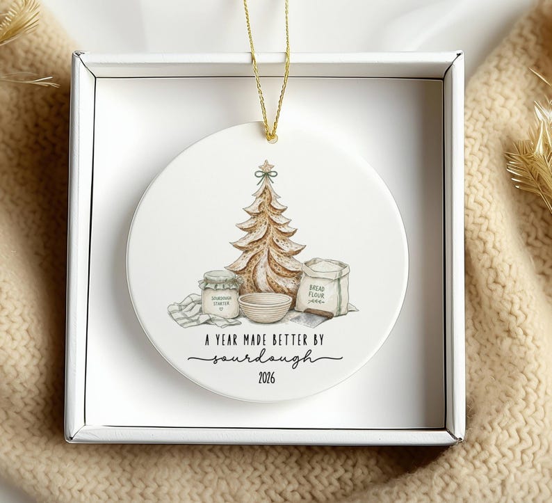 Puede incluir: Un adorno redondo de cer&aacute;mica blanca con una ilustraci&oacute;n en acuarela de un &aacute;rbol de Navidad de pan de masa madre, un frasco, un cuenco y una bolsa de harina. El texto dice A YEAR MADE BETTER BY sourdough 2026.