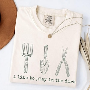 May include: Off-white t-shirt featuring a green illustration of gardening tools: a fork, trowel, and shears. The text "i like to play in the dirt" is printed below. A silver necklace with a pendant is on the shirt.