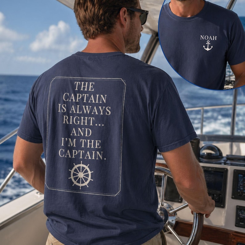 Man wearing a navy nautical captain shirt while steering a boat on the ocean. The back design reads “The Captain Is Always Right… and I’m the Captain” with a ship wheel graphic, and an inset shows the front chest design with the name Noah and an anchor.