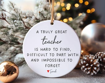 Puede incluir: Un adorno redondo de cerámica blanca que cuelga de una cinta dorada. El texto dice: A TRULY GREAT teacher IS HARD TO FIND, DIFFICULT TO PART WITH AND IMPOSSIBLE TO FORGET, seguido de un pequeño icono de corazón rojo y el nombre Sarah.